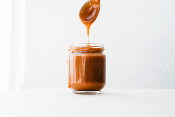 Homemade salted caramel sauce in glass jar, white background, spoon with caramel