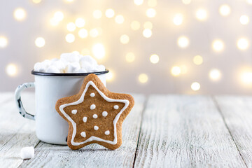 A white cup of winter hot drink with marshmallows and gingerbread star on a white wooden background with bokeh.