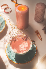Close up of pink flowers tea in blue porcelain tea cup on white table with burning candle with golden jewelry. Cozy lifestyle concept. Top view.