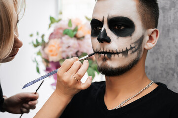 A makeup artist makes a demon makeup for a young man in a beauty salon. Halloween Celebration...