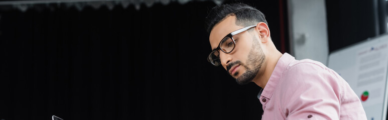 Fototapeta premium Young arabian businessman in eyeglasses looking away in office, banner