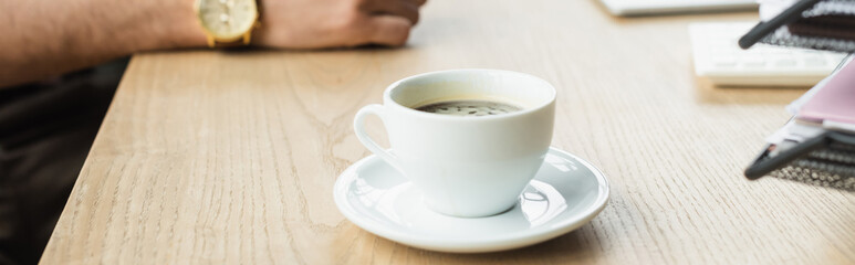 Cropped view of cup of coffee near blurred businessman in office, banner