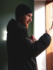 Bearded millennial man in black raincoat with smartphone in hand texting against wall with harsh shadow. Authentic male traveler Blogger guy lifestyle Solo travel adventure. Blogging communication