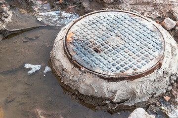 Fototapeta na wymiar A cast-iron sewer manhole surrounded by a puddle of water at a construction site. Construction of sewer wells.