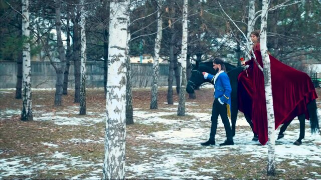 Beautiful rider woman, red velvet vintage long cloak, sits on black horse. Redhaired long hair. Fantasy fairy prince brunette man in blue tuxedo, walks holds horse by bridle. Backdrop winter nature