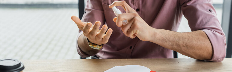 Cropped view of businessman spraying hand sanitizer near notebook in office, banner