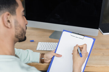 Blurred arabian businessman writing on clipboard near computer in office