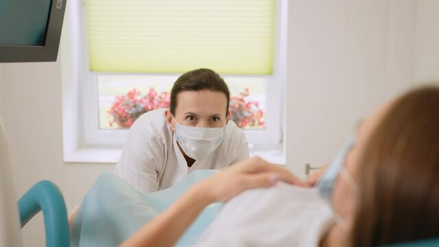 Experienced female gynecologist in lab coat, face mask and gloves holding medical vaginal speculum for examining patient. Young woman lying on gynecological chair during check up.