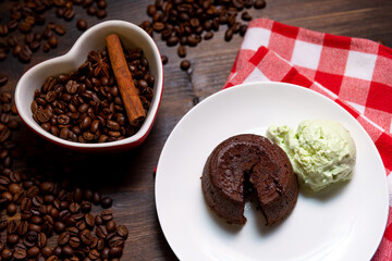 A portion of chocolate fondant with a scoop of pistachio ice cream on a plate. In the background is a heart-shaped cup filled with coffee beans and a cinnamon stick. To the right is a checkered napkin