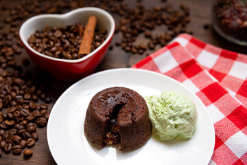 A portion of chocolate fondant with a scoop of pistachio ice cream on a plate. In the background is a heart-shaped cup filled with coffee beans and a cinnamon stick. To the right is a checkered napkin