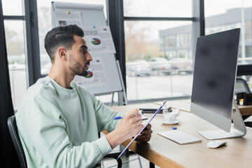 Fototapeta premium Side view of muslim businessman writing on clipboard near computer with blank screen in office