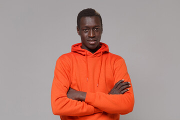 Handsome young african american man guy in orange streetwear hoodie posing isolated on grey background studio portrait. People emotions lifestyle concept. Mock up copy space. Holding hands crossed.