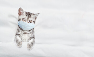 Sick kitten wearing medical protective mask lying under white warm blanket on a bed at home. Top...