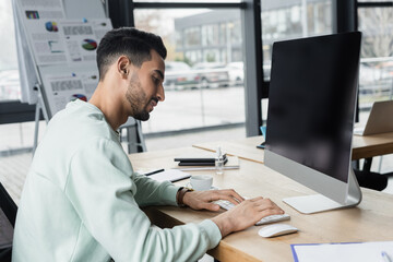 Side view of smiling arabian businessman using computer near coffee in office