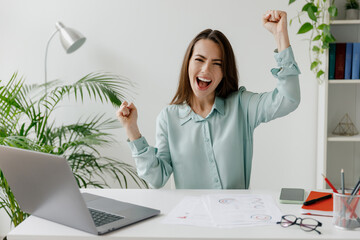 Young excited successful employee business woman in blue shirt do winner gesture sit work at...