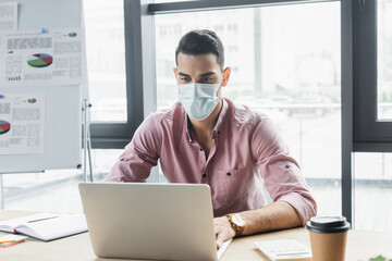 Arabian businessman in medical mask using laptop near coffee to go in office