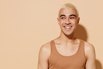 Young smiling laughing cheerful blond latin american gay man 20s with make up in beige tank shirt looking aside isolated on plain light ocher background studio portrait People lgbt lifestyle concept
