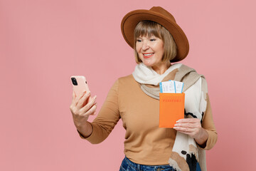Traveler tourist fun elderly senior woman 55 years old wears brown shirt hat scarf hold passport boarding tickets use mobile cell phone isolated on plain pastel light pink background studio portrait.