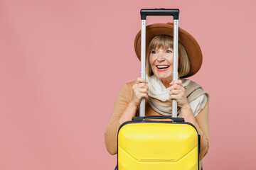 Traveler tourist fun mature elderly senior woman 55 years old wear brown shirt hat scarf hold suitcase bag hide behind handle look aside isolated on plain pastel light pink background studio portrait