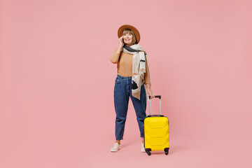 Full size traveler tourist mature elderly woman 55 years old wears casual clothes hat hold suitcase bag talk speak on mobile cell phone isolated on plain pastel light pink background studio portrait.
