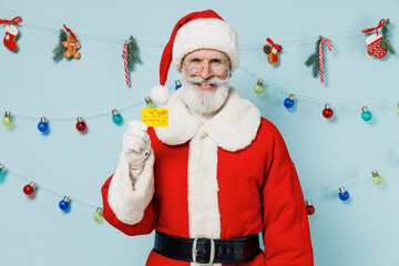 Old bearded Santa Claus man 50s in Christmas hat red suit clothes hold in hand credit bank card isolated on plain blue background studio Happy New Year 2022 celebration merry ho x-mas holiday concept