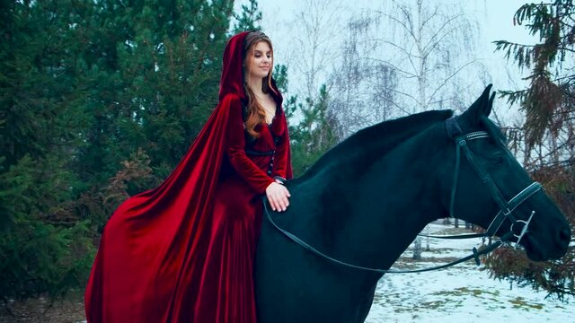 Young fantasy woman princess rider sits astride, stroking neck black horse. Enjoy winter nature green tree, beauty animal. Long velvet clothing red vintage cloak medieval cape. Hood on redhaired head