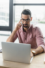 Muslim businessman in eyeglasses talking on smartphone and using laptop in office