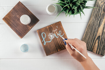 Womans hand draws word Love and dandelion flower on decorative board made of burnt wood with brush of blue paint