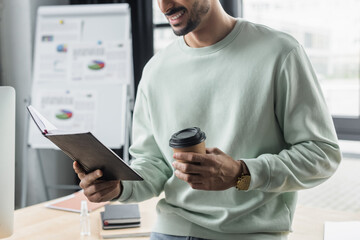 Cropped view of smiling businessman with paper cup and notebook working in office