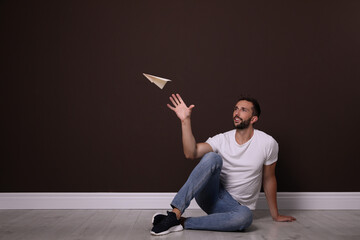Handsome man playing with paper plane near brown wall. Space for text