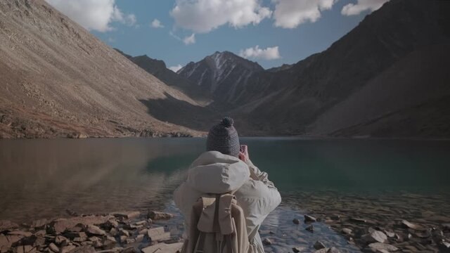 Back view of a woman in a hat with a backpack photographing a mountain landscape on the camera. Traveler by the lake on a sunny day