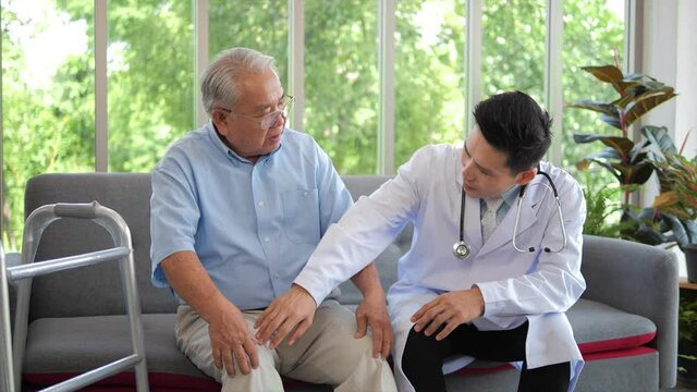 Asian male doctor wearing white lab coat checking knee of elderly man patient sitting on sofa, older people healthcare support concept.