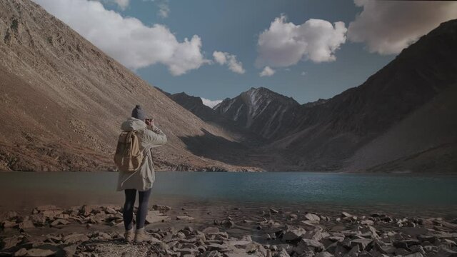 Rear view of a woman in a hat with a backpack photographing a mountain landscape on the camera. Traveler by the lake on a sunny day