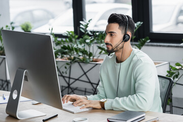 Young arabian businessman in headset using computer near cup and notebooks in office
