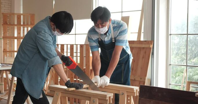 Asian man carpenter and his father in protective glasses are training woodwork apprentice how to use hand sawing in the workshop at house