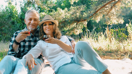 A middle-aged couple with glasses of wine is having fun chatting online while sitting in nature on a picnic on a summer evening.