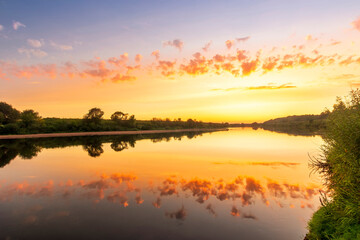 Scenic view at beautiful summer river sunset with reflection on water with green bushes, grass, calm water ,deep cloudy sky and glow on a background, spring , evening landscape