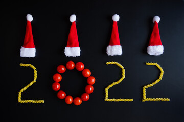 New year 2022 numbers. Happy new year symbol. Holidays creative concept. Healthy vegetarian food. Fresh red tomatoes, pasta,  Christmas hats on black background. Christmas greeting card. Copy space.