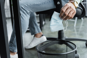 Cropped view of businessman holding medical mask near trash can in office