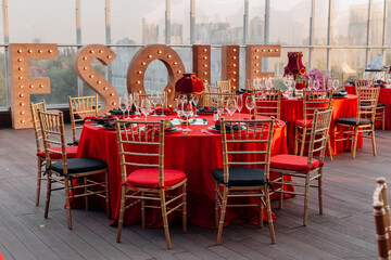 Guest's tables and chairs arrangement for banquet in black, red and gold style. Elegant event hall: decor, tablecloth, plates, napkins, roses, glasses. Themed party celebration on the roof, outdoor.