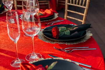 Guest table setting for banquet in black, red and gold style. Elegant and luxury dinner arrangement: decor, tablecloth, plates with napkins and fresh roses, glasses, cutlery. Themed party celebration