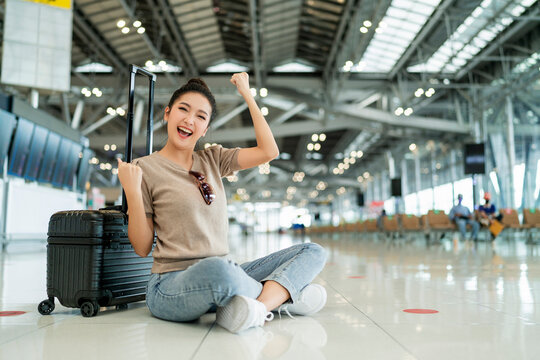 lockdown is over time to travel,happiness asian femlae traveller wear casual cloth hand wave gesture smiling while sit relax on terminal airport floor with luggage safety travel concept