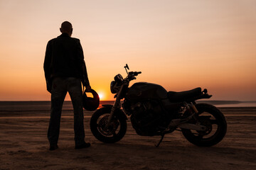 Fototapeta na wymiar Bold senior man wearing leather jacket posing with motorcycle outdoors
