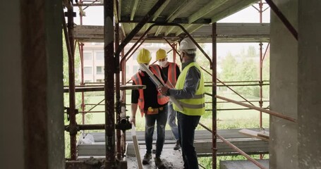 People working in construction site wearing face mask during Covid pandemic. Men at work in new house inside building. Two workers talking with engineer, looking at blueprint during business meeting - Powered by Adobe