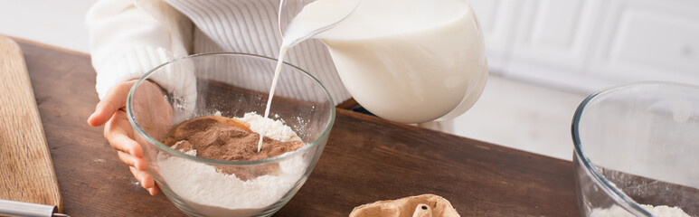 Cropped view of african american woman pouring milk in flour with cocoa while cooking in kitchen, banner