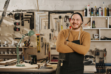 Smiling carpenter in a watch cap standing with hands crossed in a big workshop