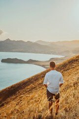 Naklejka na ściany i meble A young man travels along the mountain coast at sunset. Rear view A man stands on a mountainside and enjoys the view. The concept of freedom and achievement of goals.