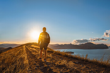 A young male tourist with a backpack walks along the path to the top of the mountain at sunset. Goals and achievements concept