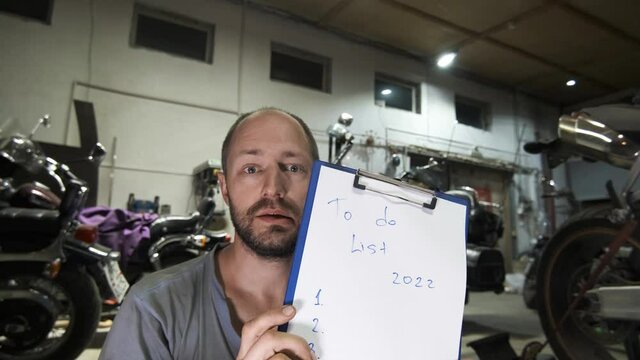 A mechanic guy in a blue short-beard T-shirt stands next to a motorcycle, shows an unfilled 5-point to-do list for 2022, and talks while gesturing in front of a motorcycle workshop