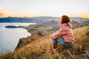 A beautiful young woman tourist is sitting on top of a mountain and enjoying the view of the sunset sea and mountains. The concept of freedom and meditation, an active lifestyle.